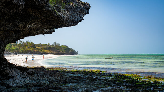 View of clear turquoise waters gently lap against the white sandy beach, shadowed by rugged rock formations, inviting exploration in Paje, Unguja South Region, Tanzania.