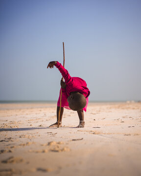 Paje, Tanzania - 23 January 2025: View of a child in vibrant pink bending low on the sandy beach, a slender stick mirroring the curve of their form against the vast, serene sky.