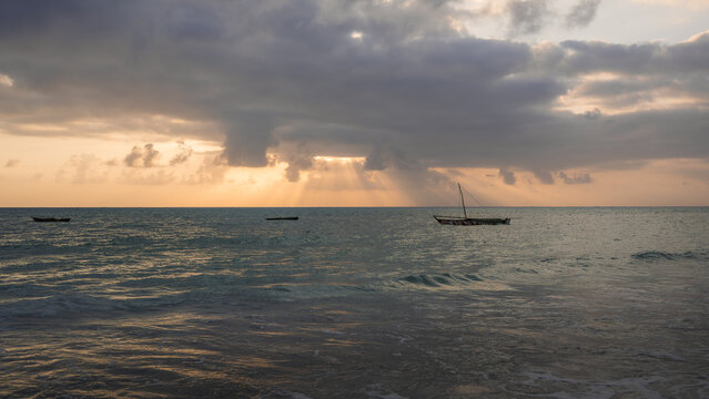 View of the tranquil turquoise waters reflecting the golden sunset, with traditional boats silhouetted against the horizon under a dramatic sky, Paje, Tanzania.