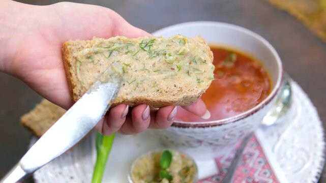 spread garlic lard with herbs onto slices of bread using a knife. Close-up of a traditional Ukrainian appetizer served in a rustic outdoor setting with a wooden table and embroidered cloth.