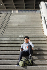 Male sitting on wide concrete staircase in sunlight with hoodie headphones smartphone green duffel © wavebreak3