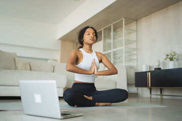 Obraz premium Yoga mindfulness meditation online. African woman practicing yoga with online lessons in laptop at home. Woman sitting in lotus pose meditating relaxing. Girl doing breathing practice. Yoga at home