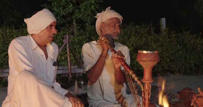 Indian old aged man wear white kurta pajama sitting on woven cot bed enjoy desi life at rural area. Happy two male group talk speak share gossip do hookah hold pipe spend night time village place