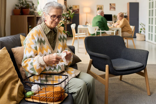 Senior Caucasian woman knitting while sitting in retirement home lounge, supporting active aging and hobby therapy. Useful for senior care, assisted living, wellness marketing