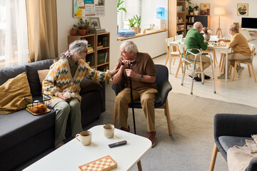 Wide shot of senior residents socializing in care home, woman comforting man with cane while couple...