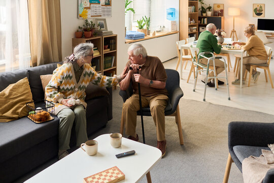Wide shot of senior residents socializing in care home, woman comforting man with cane while couple playing board game in background. Ideal for elder care, assisted living, support services