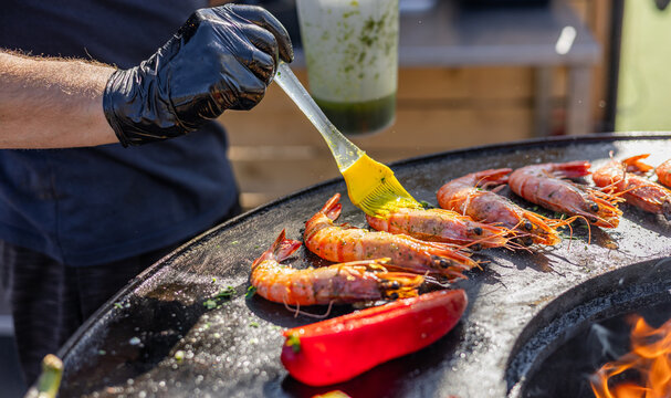 Variety of food being cooked on a grill. The food includes shrimp, corn on the cob, and flatbreads, which are all being cooked over an open fire. The food is being cooked in a round metal pan.