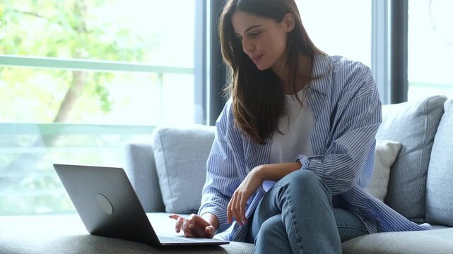 Happy Young Woman Smiling While Comfortably Working on Her Laptop Computer from Home Sofa.