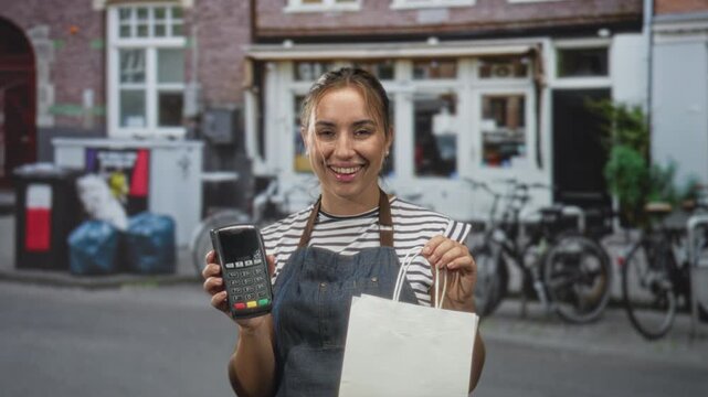 Woman in apron offering a dataphone with hand while holding a paper bag and smiling at customer on street; friendly service.