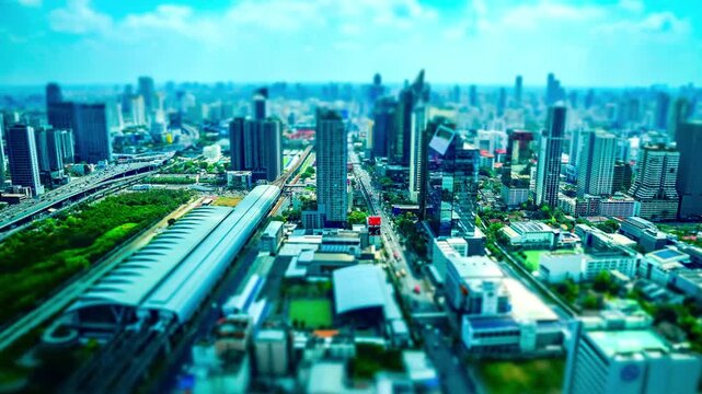 A tilt-shift style aerial view of the city&rsquo;s modern skyline, with dense high-rises, elevated roads, and a green transport corridor compressed into a miniature city effect beneath a bright cyan sky.