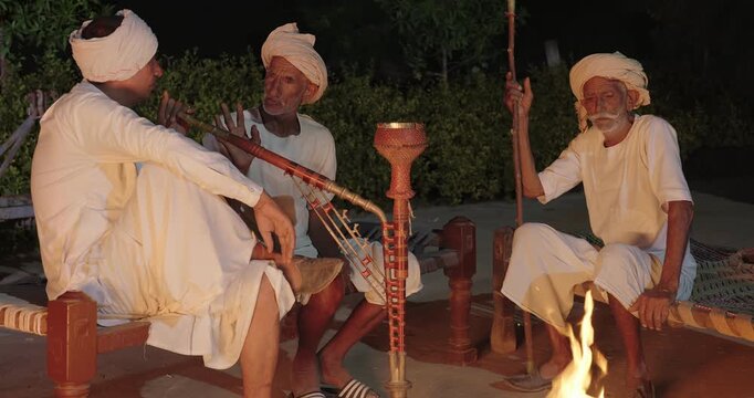 Indian old aged man wear white kurta pajama sitting on woven cot bed enjoy desi life at rural area. Happy three male group talk speak share gossip do hookah hold pipe spend night time village place
