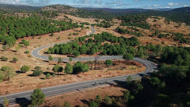 Drone pulling back over winding road in Madrid countryside with cyclists riding through dry landscape and hills.