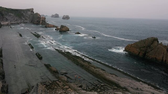 Drone panning along La Arn&iacute;a beach in Cantabria showing flysch rock formations, waves and rugged coastline under cloudy sky.