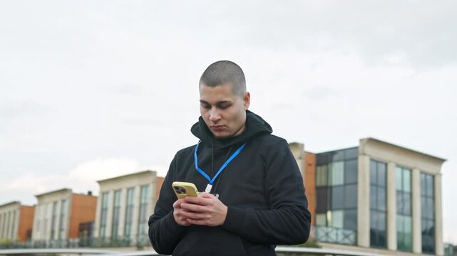 Young employee with a buzz cut and lanyard using his smartphone while standing outdoors in front of an office building