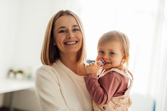 Mother holding smiling daughter with pacifier indoors in daylight