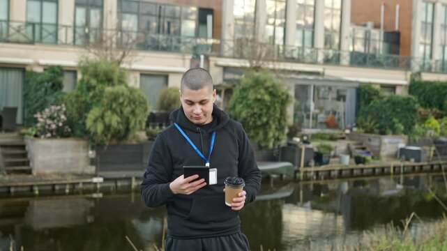Young employee using tablet and drinking coffee outdoors during a break next to a canal in a modern neighborhood