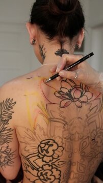 Close-up of a female tattoo artist draws a scketch of intricate floral and mandala design on a woman's upper back. Free hand tattoo technique.