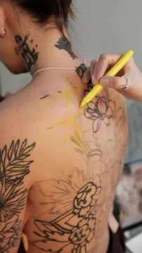 Close-up of a female tattoo artist draws a scketch of intricate floral and mandala design on a woman's upper back. Free hand tattoo technique.