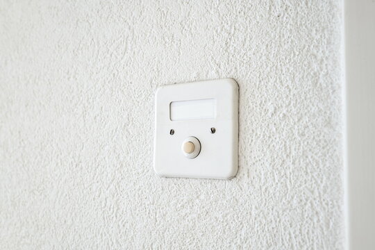 Close up of a classic square doorbell with a round button and name plate on a rough white wall