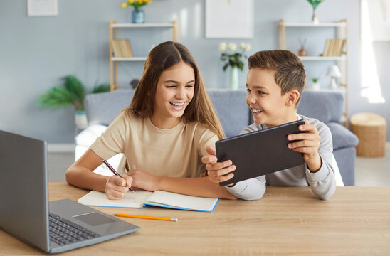 Two children are happily doing their homework and having fun using modern gadgets. Girl is sitting in front of laptop and writing in notebook, while boy shows her what is displayed on screen of tablet