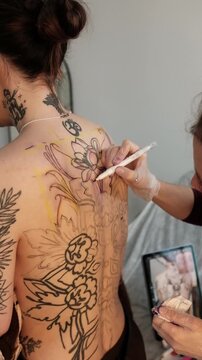 Close-up of a female tattoo artist draws a scketch of intricate floral and mandala design on a woman's upper back. Free hand tattoo technique.