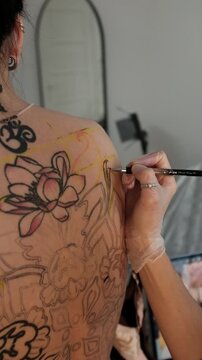 Close-up of a female tattoo artist draws a scketch of intricate floral and mandala design on a woman's upper back. Free hand tattoo technique.