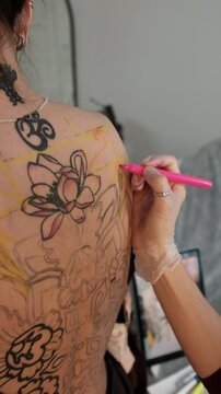 Close-up of a female tattoo artist draws a scketch of intricate floral and mandala design on a woman's upper back. Free hand tattoo technique.