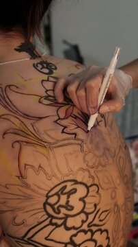 Close-up of a female tattoo artist draws a scketch of intricate floral and mandala design on a woman's upper back. Free hand tattoo technique.
