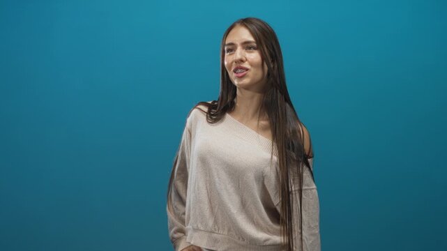 Young woman with braces smiling, long hair over one shoulder, wearing a sweater, posed against a teal studio backdrop; confidence youth.