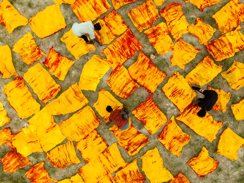 Narayanganj, Bangladesh - 30 March 2026: Aerial view of vibrant yellow fabrics, patterned with fiery red, drying under the sun, as workers tend to the textile spread.