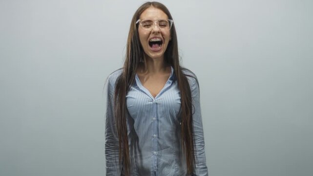 Teenage girl with braces and clear glasses screams with mouth wide open showing tongue, long hair and striped button shirt in a plain studio; excitement youth.