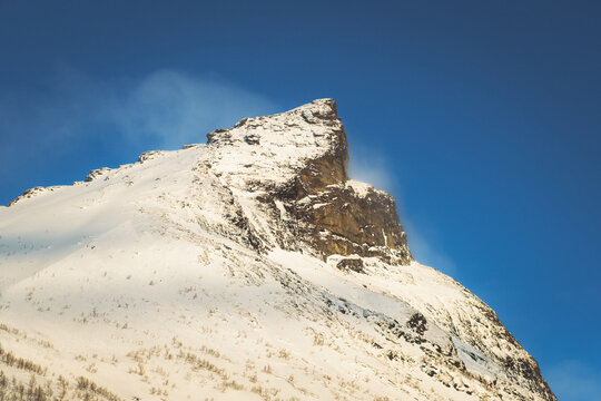 View of a snow-dusted mountain peak rises majestically against a clear, vibrant blue sky, a contrast of cold whites and warm stone textures, Indregard, Troms, Norway.