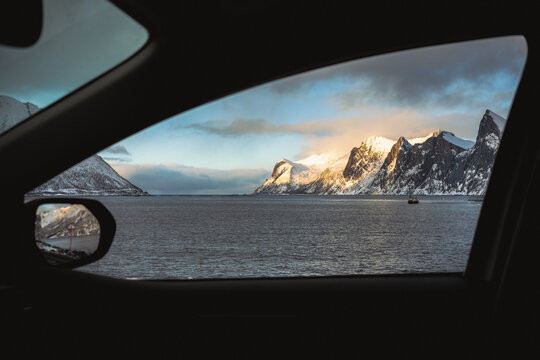 View of the sea meeting snow-capped mountains under a cloudy sky, framed by the dark interior of a car, capturing the essence of a journey, Indregard, Troms, Norway.