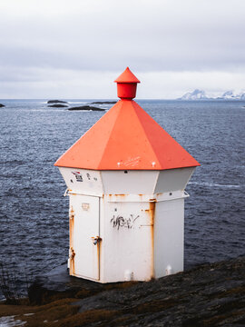 View of a white, rust-stained structure with a vivid orange roof stands resilient against the backdrop of the moody ocean and distant mountains, Henningsvaer, Norway.