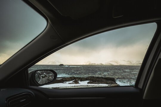 View of choppy ocean waters meet rugged, snow-dusted islands under a cloudy sky, framed by a car window's dark interior, Henningsvaer, Nordland, Norway.