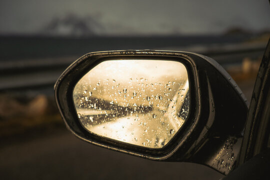 View of raindrops cling to a side mirror reflecting the distant, moody coastline under a muted sky, creating a blurred scene, Henningsvaer, Nordland, Norway.
