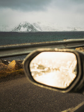 View of snow-capped mountains reflecting in a car's side mirror, juxtaposed against the dark ocean water under a cloudy sky, Henningsvaer, Norway.