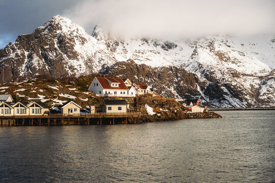 View of white houses with red roofs nestled along the coast, contrasting with the snow-capped mountain backdrop and calm waters, Henningsvaer, Nordland, Norway.