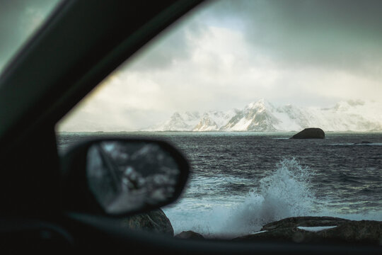 View of the rugged, snow-dusted mountains beyond the turbulent, slate-gray sea, framed by the car's interior and mirror reflecting the wild scene, Henningsvaer, Norway.
