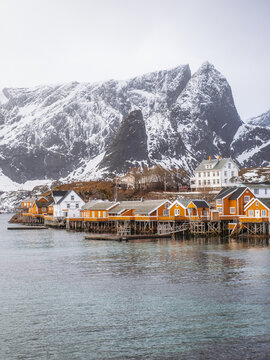 View of quaint yellow and white stilt houses nestled along the tranquil waters, backed by snow-dusted mountains piercing the sky, Hamnoy, Nordland, Norway.