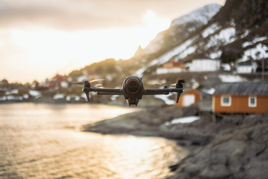 View of a drone hovering above the water as the sun sets behind snowy mountains and orange houses in Tind, Nordland, Norway.