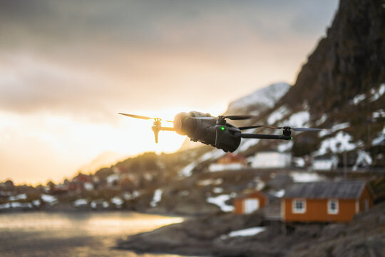 View of a drone in flight over the serene Lofoten Islands, with snow-capped mountains, rustic cabins, and a tranquil body of water, Tind, Nordland, Norway.