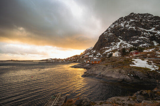 View of golden light kissing the tranquil waters near rocky shores, contrasting with snow-dusted peaks and quaint red-roofed houses, Tind, Nordland, Norway.