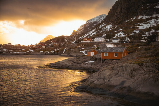 View of the orange houses nestled on rocky shores, their windows glowing warmly against the backdrop of snow-dusted mountains and golden sunset , Tind, Nordland, Norway.
