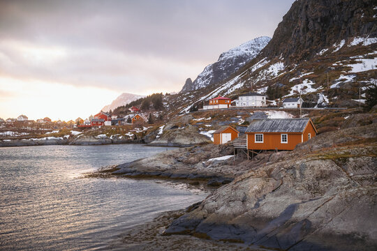 View of rustic red houses nestled on rocky shores under snow-dusted mountains, where the sea meets the sky in Tind, Nordland, Norway.