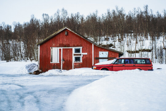 View of a red barn stands boldly against the stark white snow, a matching red car parked nearby, under the watchful gaze of bare trees, Bardufoss, Troms, Norway.