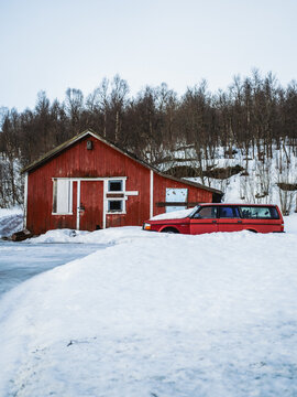View of a vibrant red vintage car parked in deep snow near a rustic red wooden building, creating a striking contrast against the white landscape, Bardufoss, Norway.