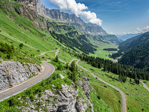 Aerial view of winding roads carve through verdant valleys beneath towering cliffs, a symphony of green and grey, Spiringen, Uri, Switzerland.