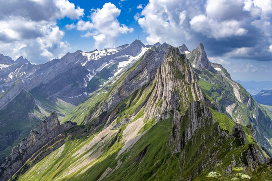 Aerial view of verdant slopes meeting jagged peaks capped with snow under a vibrant sky, Schwende-Rute, Appenzell Innerrhoden, Switzerland.