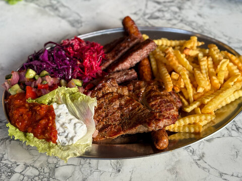 Plate with assorted grilled meat, steak, and cevapcici, served with mixed salad, sauce and French fries high-angle view, traditional Balkan food.